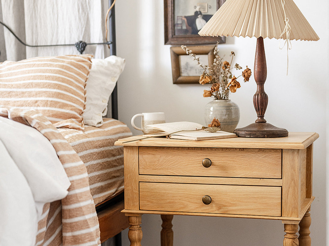 A bedside table with a lamp and vase next to a bed dressed with linen sheets
